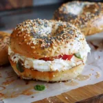 Everything bagel topped with cream cheese, tomato slices, and fresh basil, served on parchment paper.